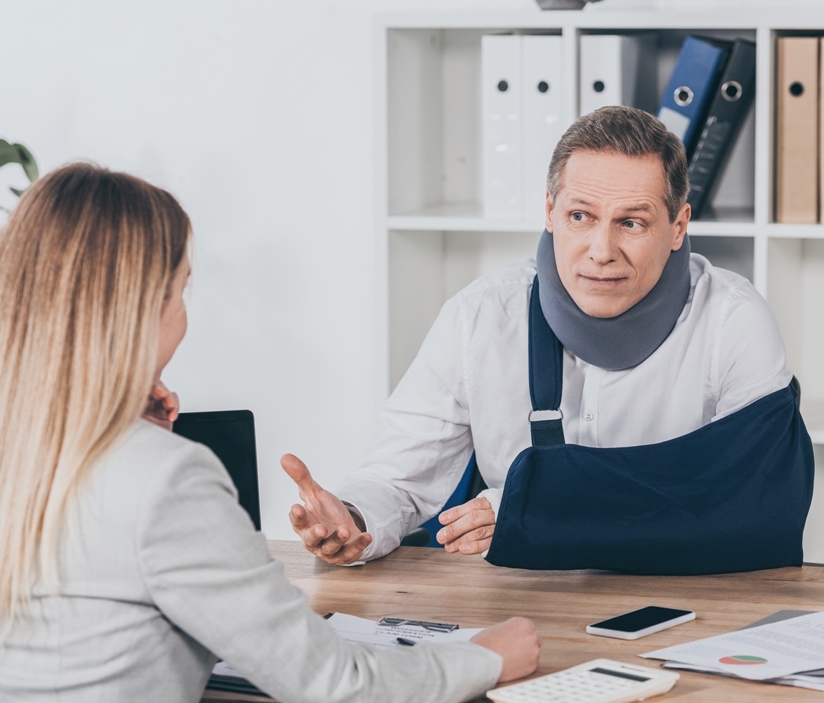 middle aged worker in neck brace with broken arm sitting at table and talking to blond woman in